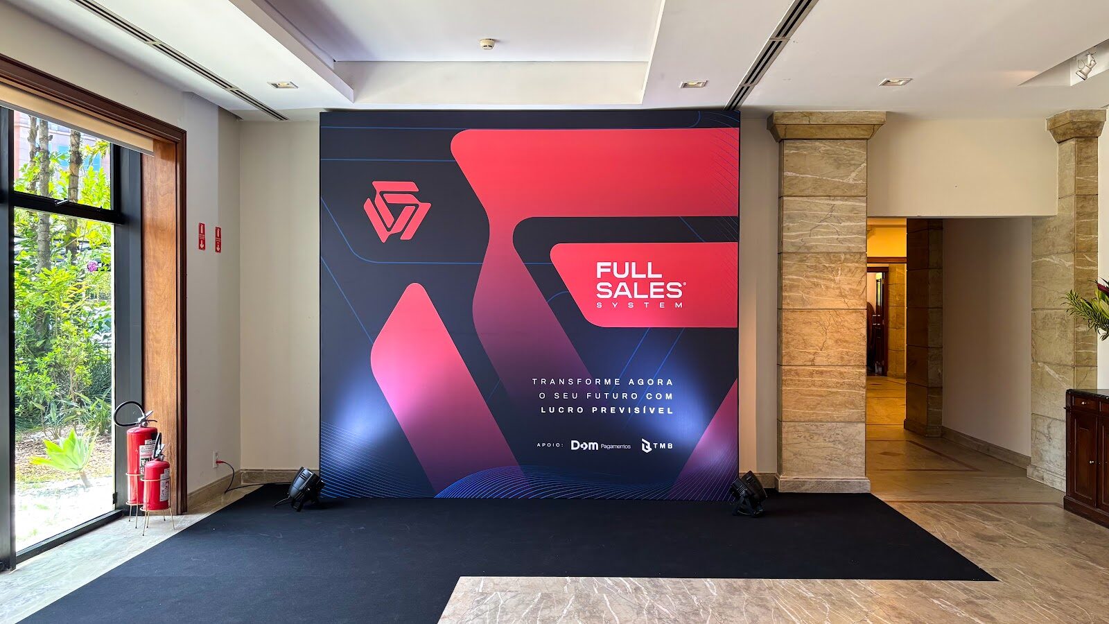 Backdrop cenográfico vermelho e preto montado sobre carpete no lobby de mármore de um evento corporativo