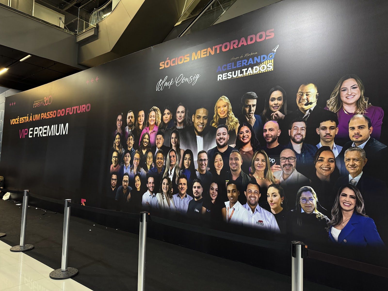 Painel cenográfico preto com fotos de palestrantes e totens de isolamento em evento corporativo