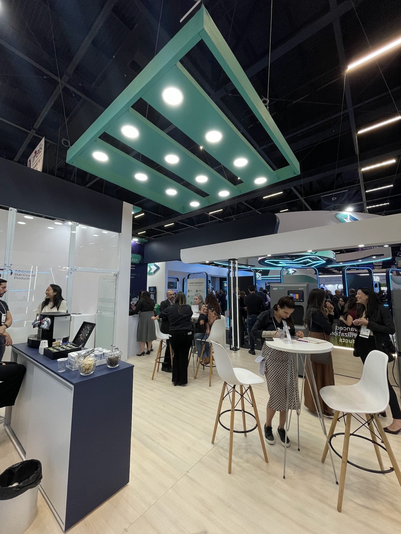 Lounge interno de stand com estrutura triangular suspensa iluminada e mesas altas