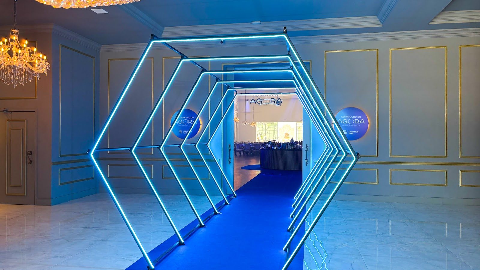 Túnel cenográfico hexagonal com neon azul formando corredor de entrada em evento corporativo