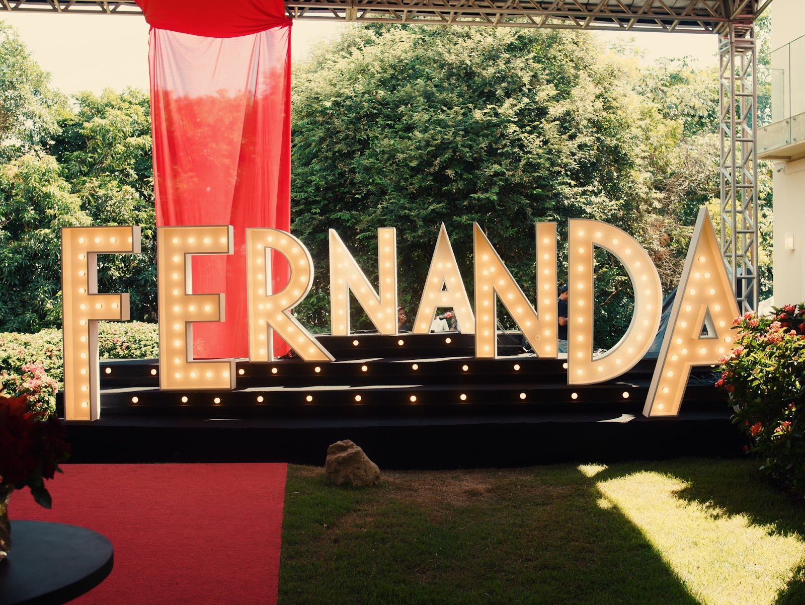 Letreiro luminoso em jardim com tapete vermelho e estrutura treliçada para evento temático