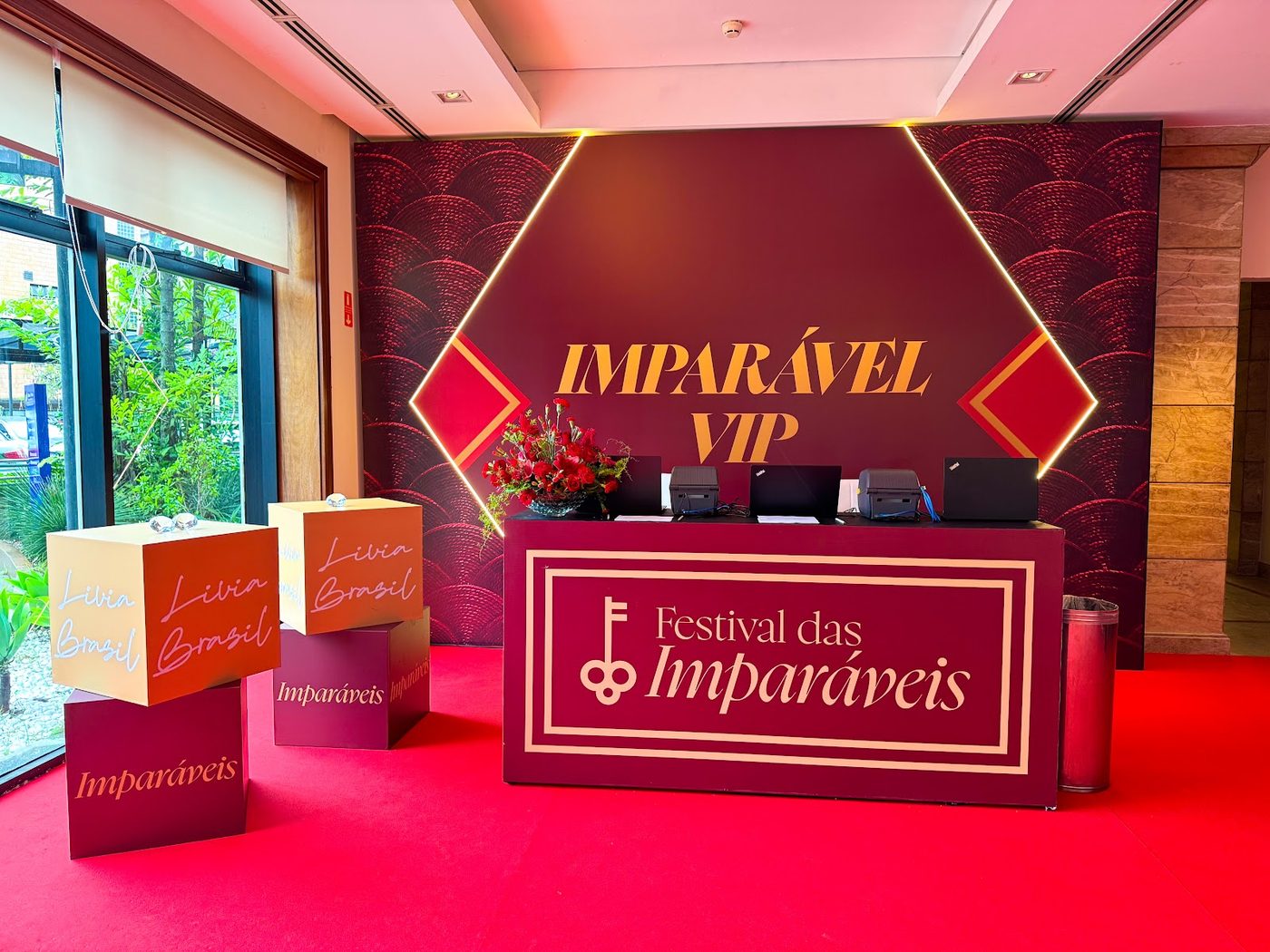 Backdrop cenográfico de check-in com tons de vinho e dourado e arranjo floral — cenografia temática para evento corporativo