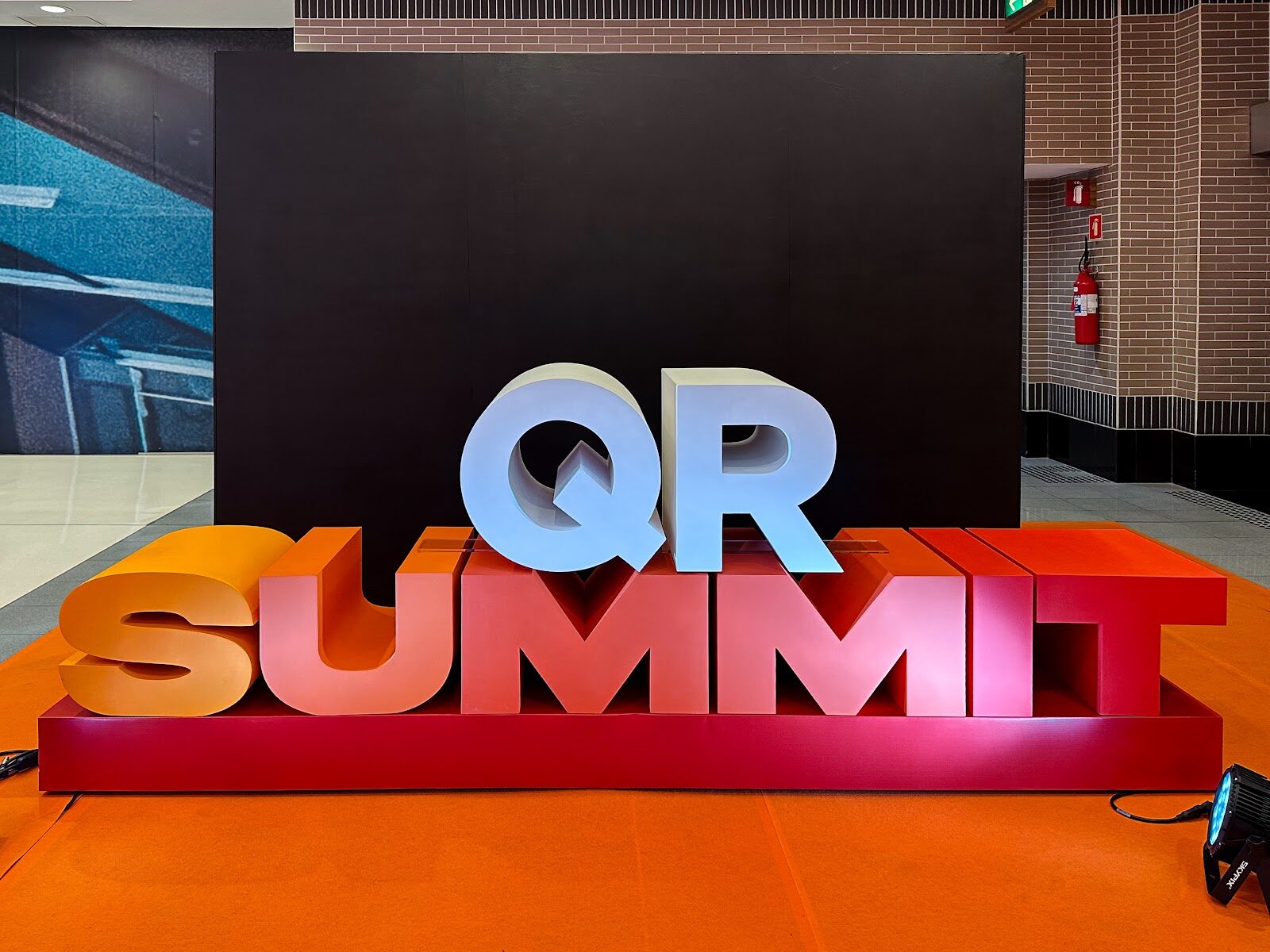 Escultura cenográfica tridimensional com letras QR Summit sobre tapete laranja