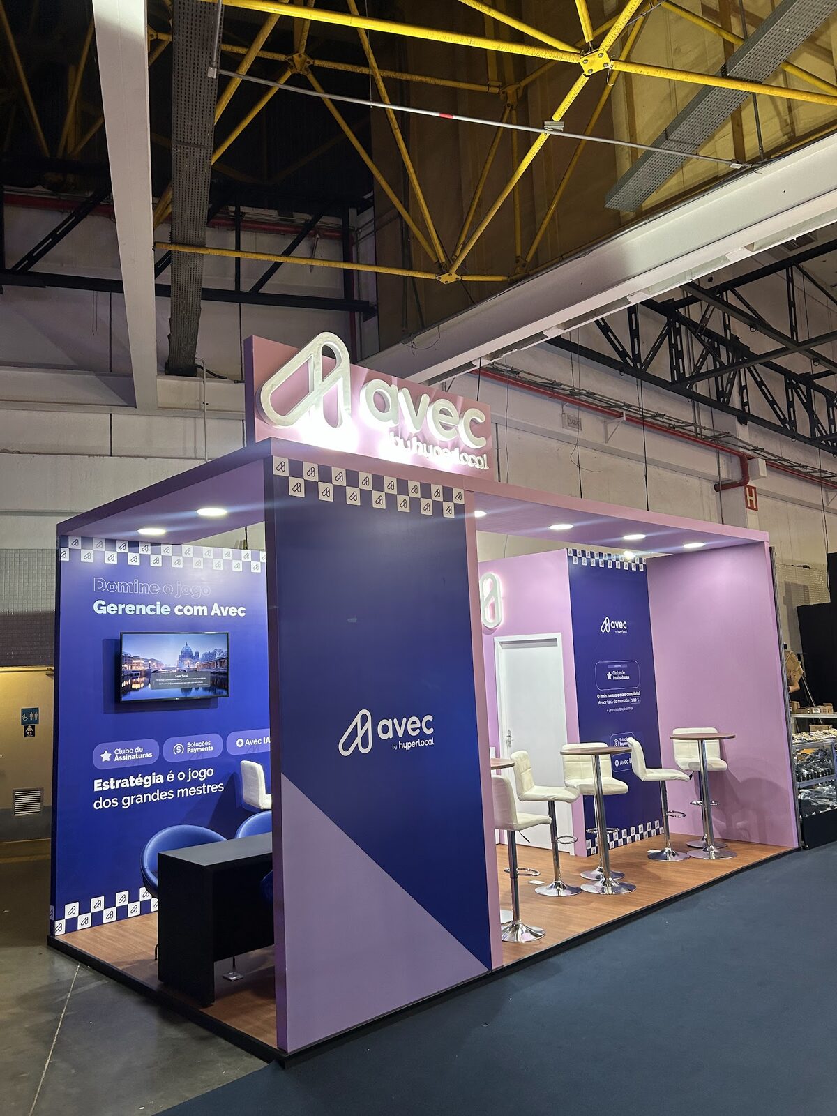 Stand da Avec no Expo Center Norte com iluminação linear, telas de vídeo e a estrutura metálica do pavilhão ao fundo