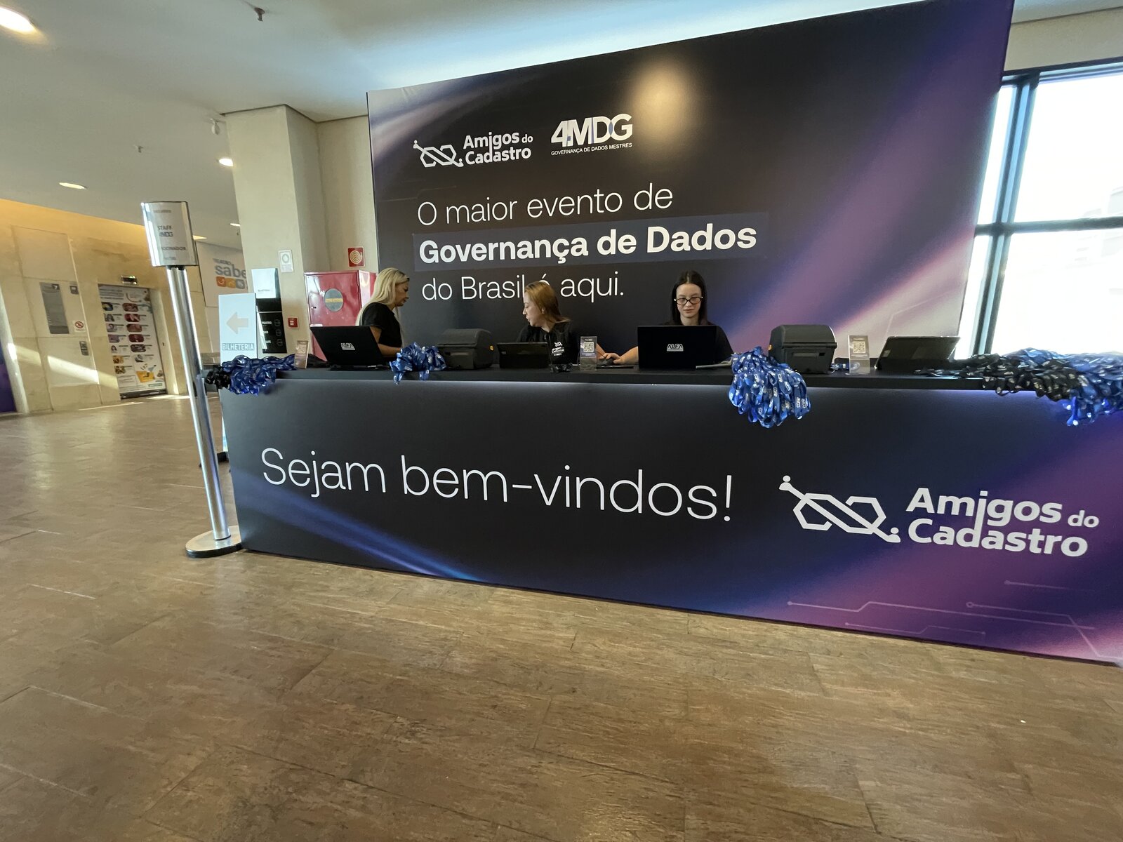 Credenciamento do evento Amigos do Cadastro com backdrop de marca e balcão iluminado por fita LED azul, produzido pela Atom Cenografia