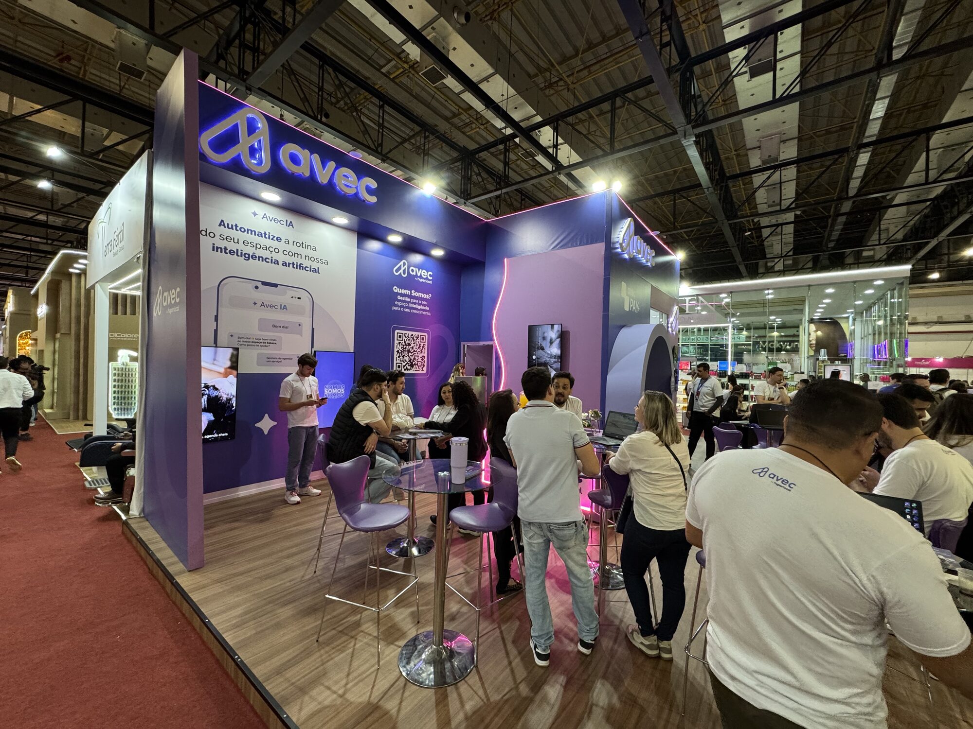 Área de relacionamento do stand da Avec na Beauty Fair 2025, com mesas bistrô e telas de demonstração
