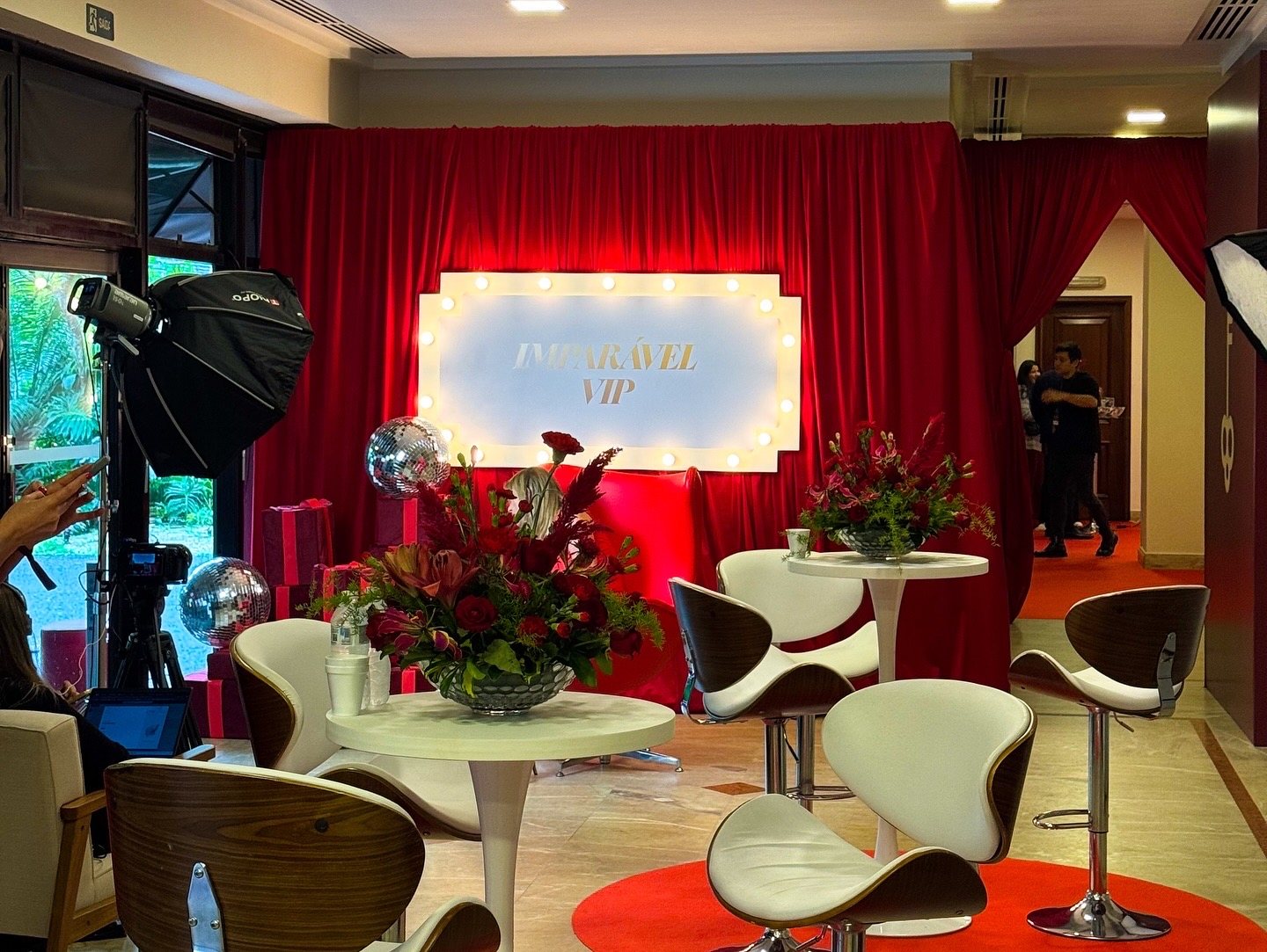 Lounge VIP com cortina vermelha, placa luminosa IMPARÁVEL VIP em filamento, mesas e banquetas e composições com flores